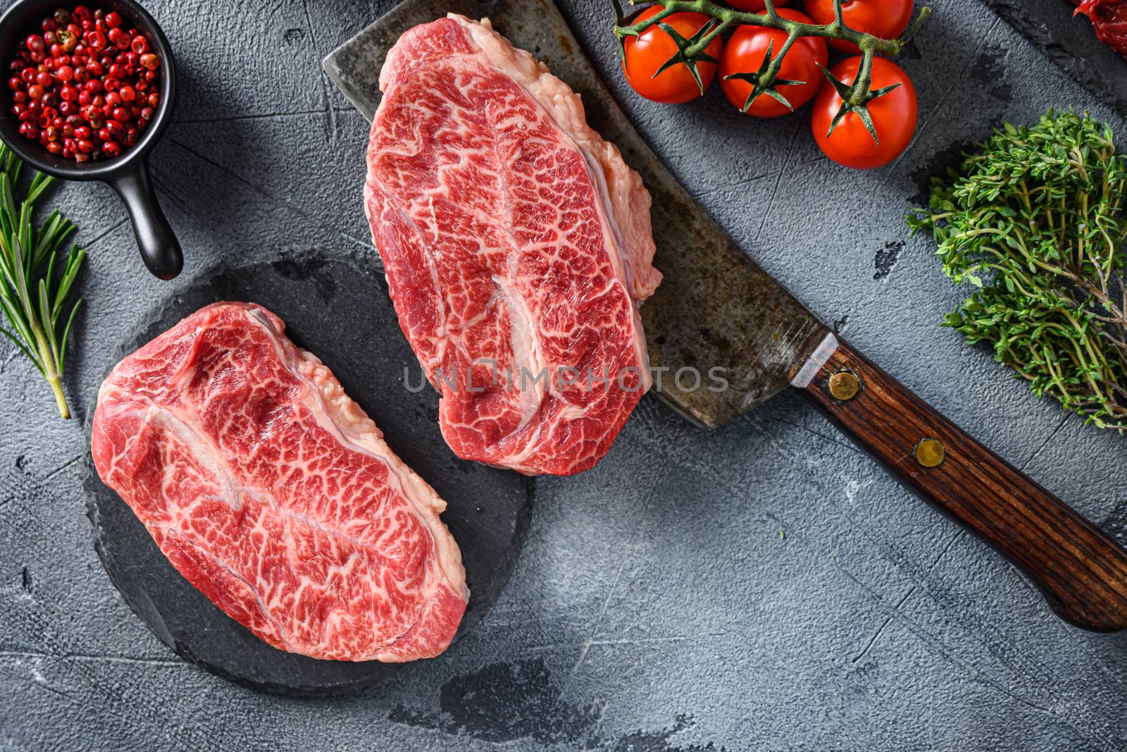 Raw top blade oyster Steak on stone and meat butcher cleaver, marbled beef with herbs tomatoes peppercorns top view.