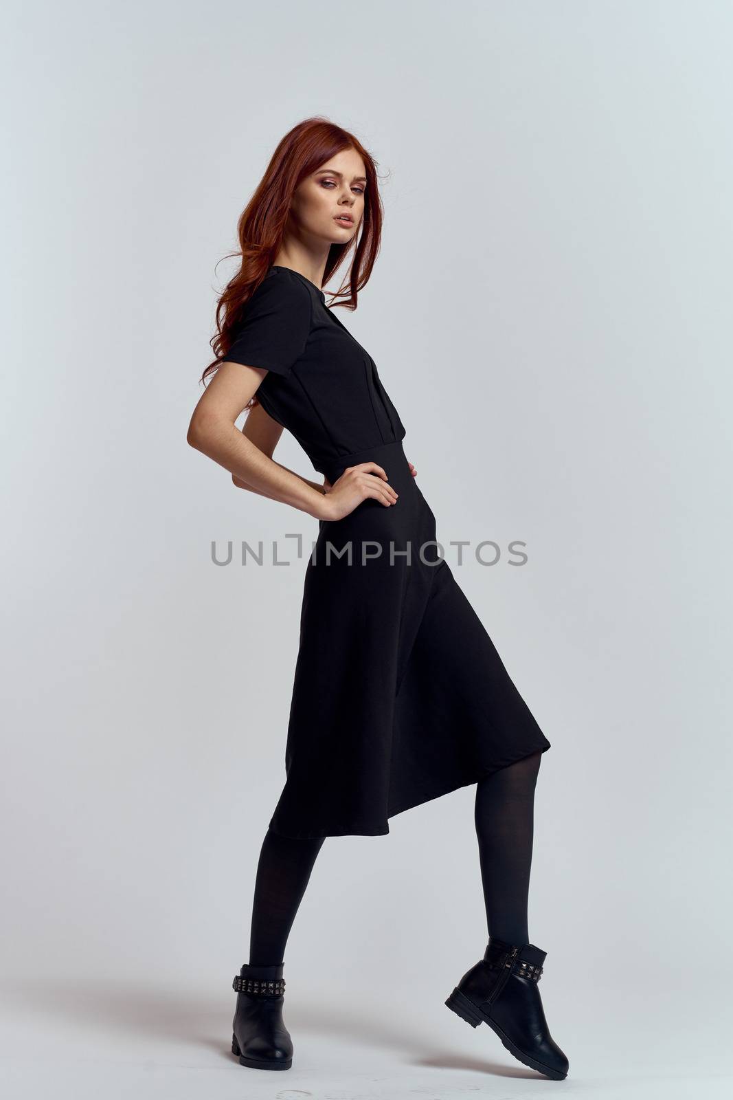 A woman in a black dress on a light background and pantyhose shoes red hair and pose in full growth. High quality photo