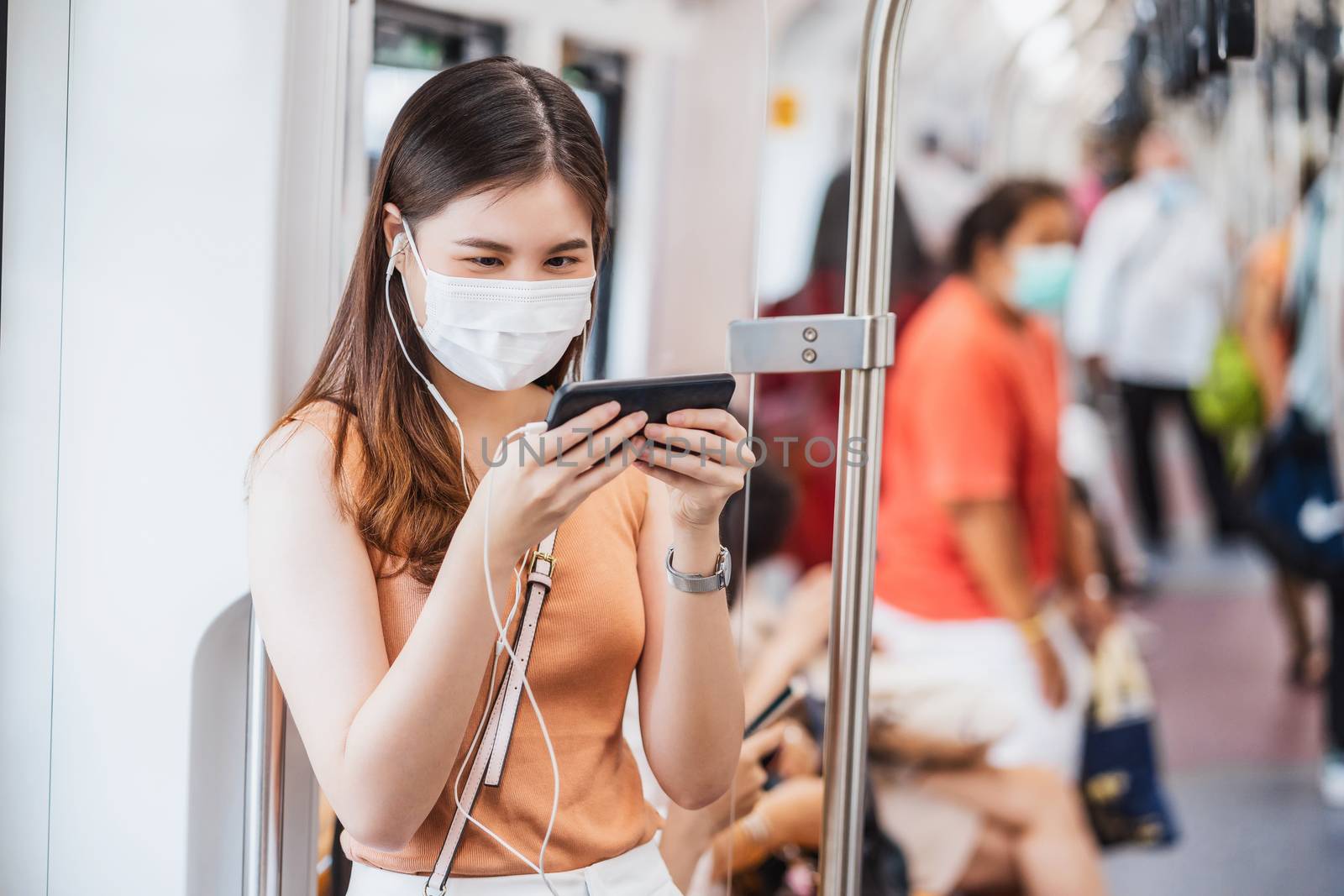 Young Asian woman passenger wearing surgical mask and listening music via smart mobile phone in subway train when traveling in big city at Covid19 outbreak, Infection and Pandemic concept