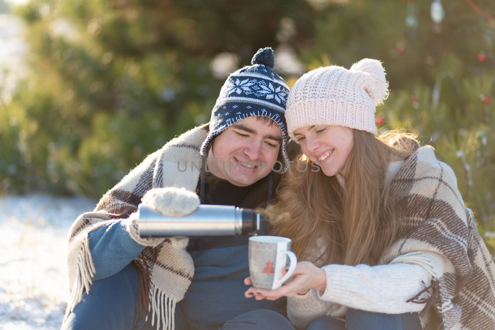 Young couple in love drink a hot drink from a thermos, sitting in the winter in the forest, tucked into warm, comfortable rugs, and enjoy nature. The guy pours a drink from a thermos in a cup by Try_my_best