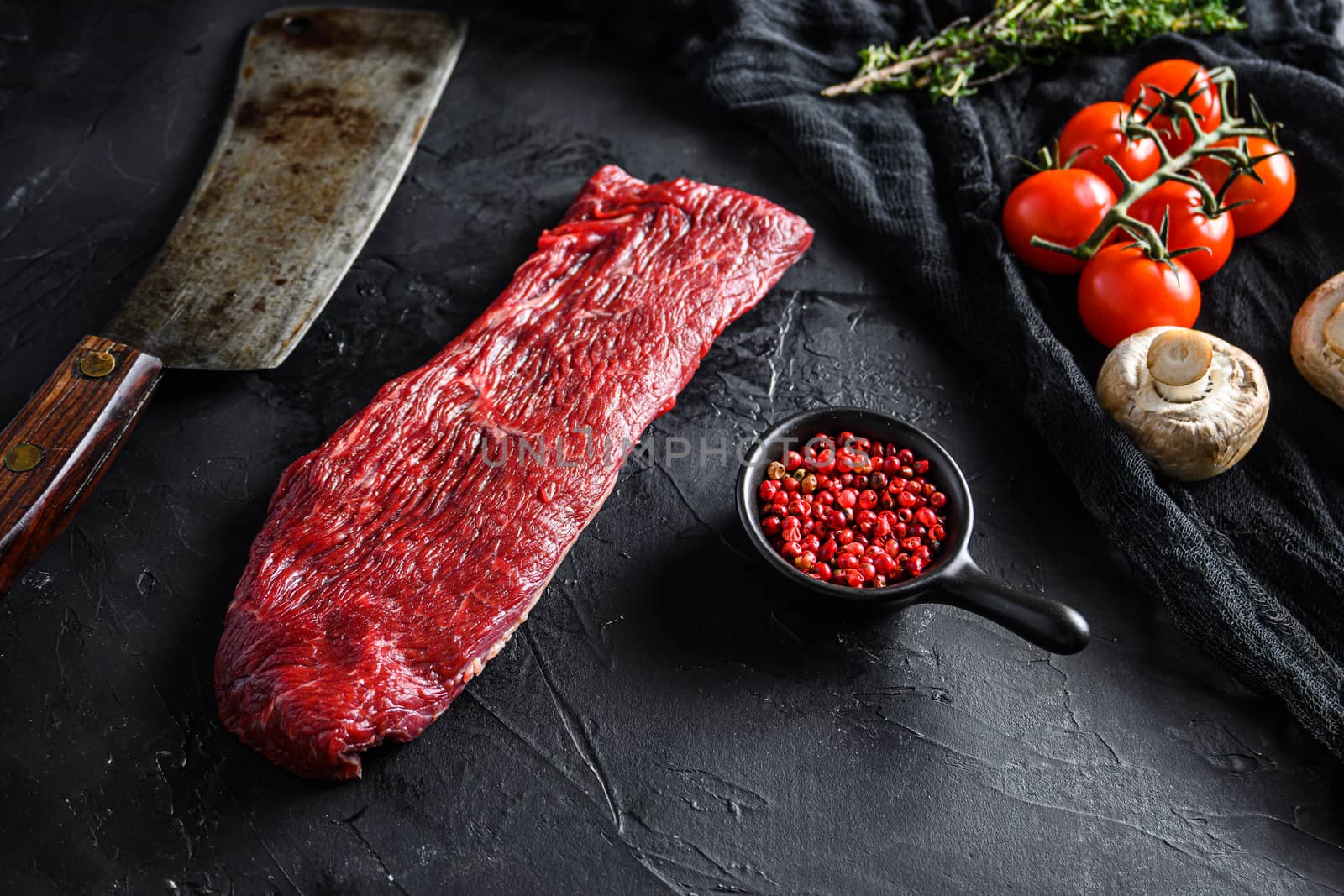 Raw triangle roast or tri tip, near butcher knife with pink pepper and rosemary. Black background. side view.