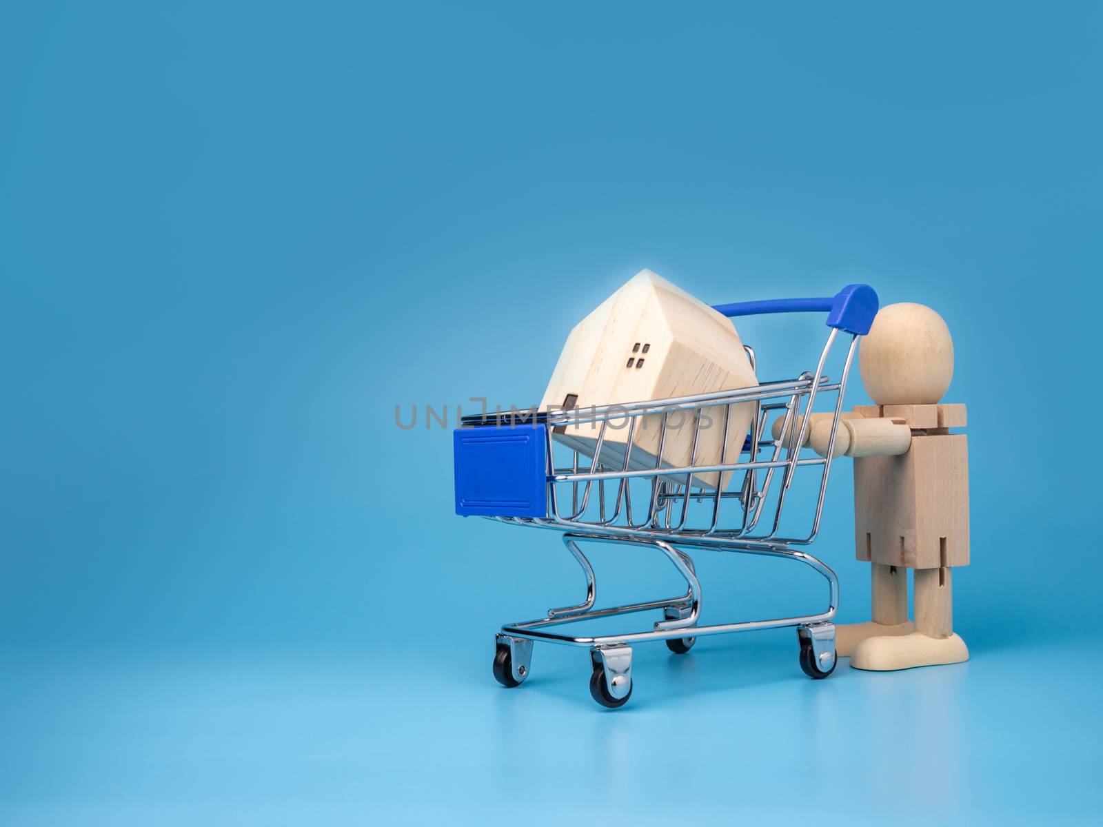 Wooden dolls that Stand next to the shopping cart With a model wooden house on top And has a blue background. Home buying and selling ideas