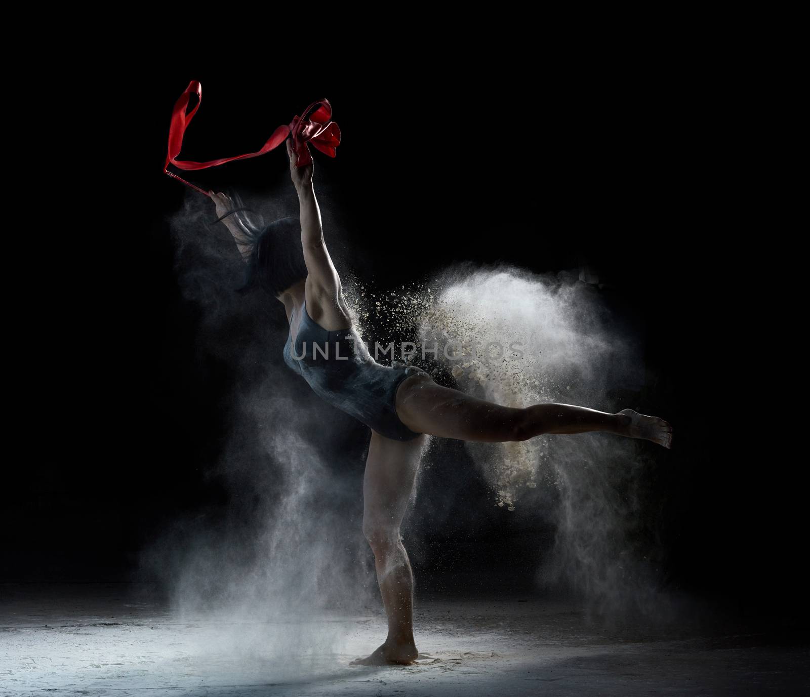 beautiful caucasian woman in a black bodysuit with a sports figure dancing with red ribbon on a black background, gymnastic exercises in flying white flour