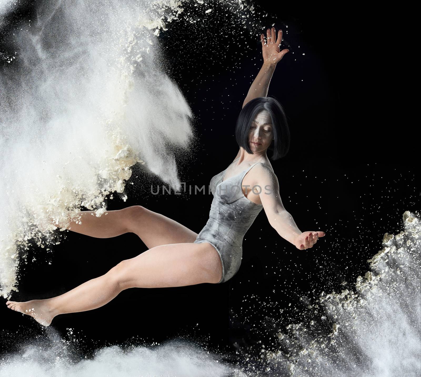 beautiful young caucasian woman in a black bodysuit with a sports figure soars on a black background in a spray of white flour, creative photo, levitation effect