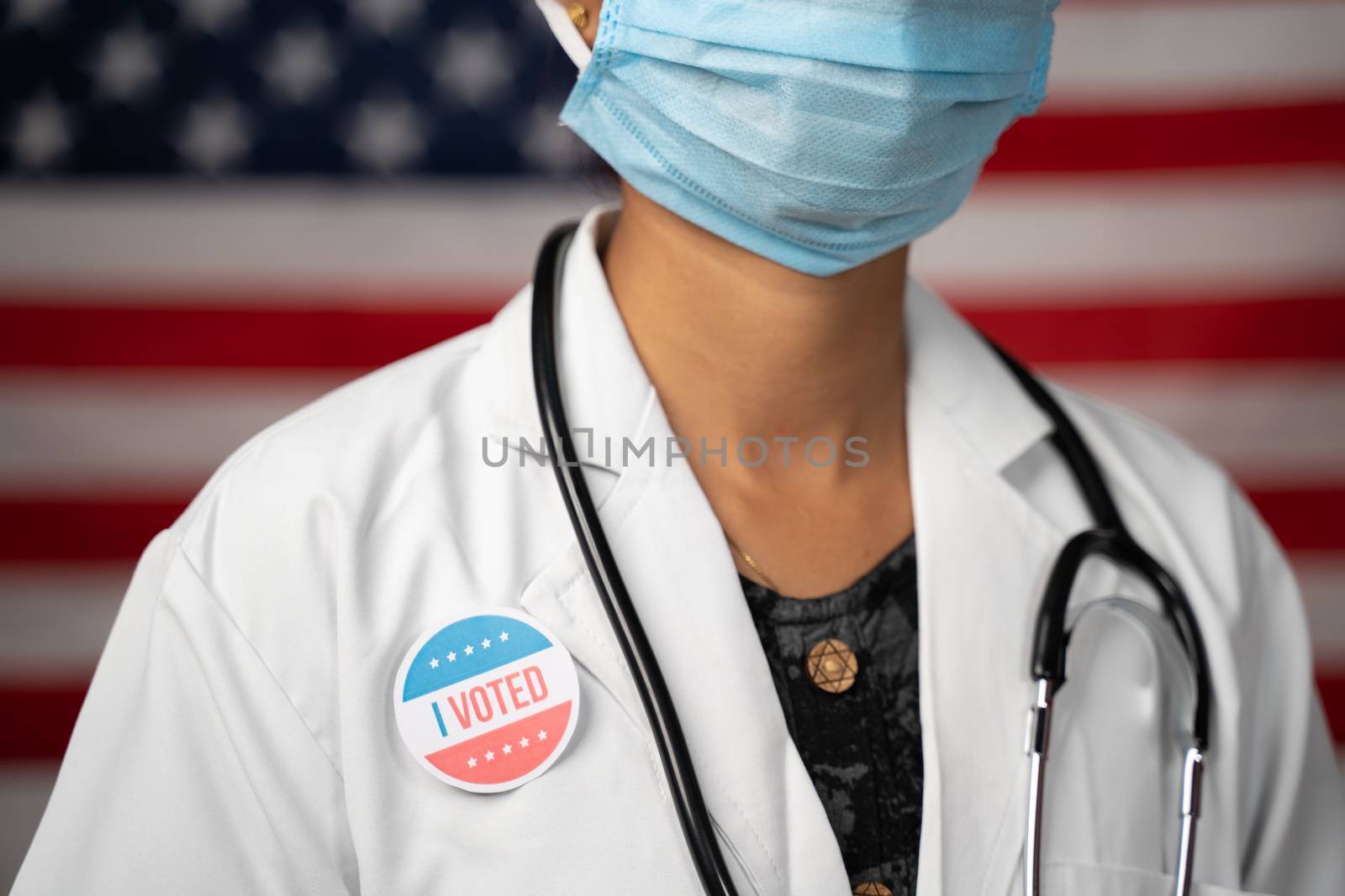 Doctor or nurse medical mask and stethoscope wearing placing I voted sticker to her apron - concept of US election with US flag as background