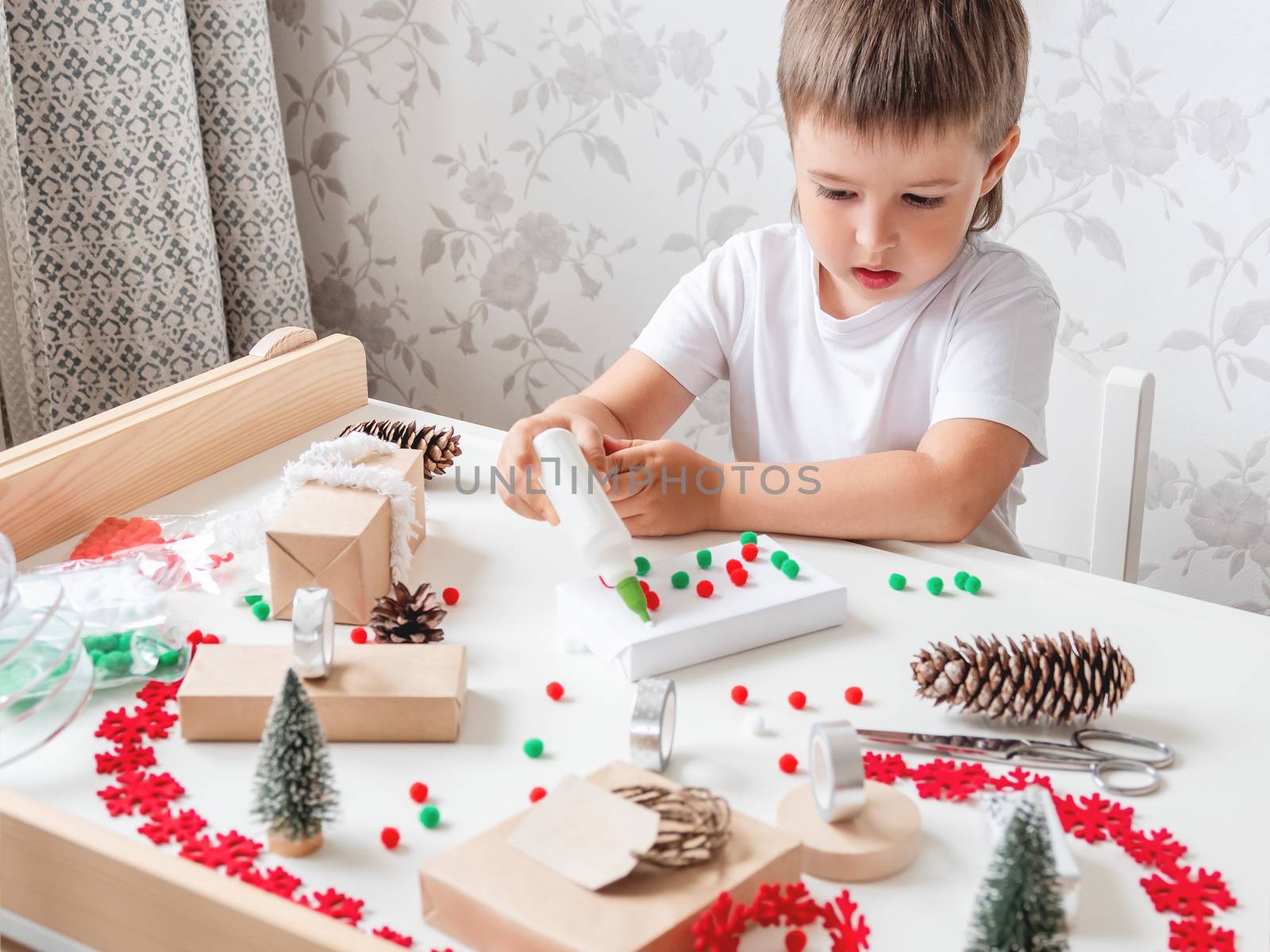 Kid wraps handmade Christmas presents in craft paper with colorful pompons and snowflake ribbons. Child prepares gifts for New Year celebration. Peaceful leisure activity before winter holidays.