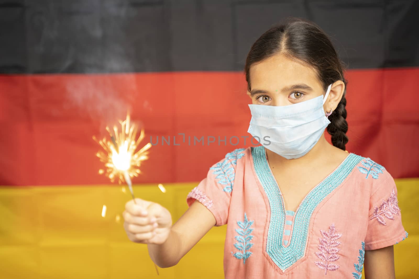 Young Girl Kid on medical mask holding Sparkler with german flag as background - concept showing Celebration of German Unity or Republic Day.
