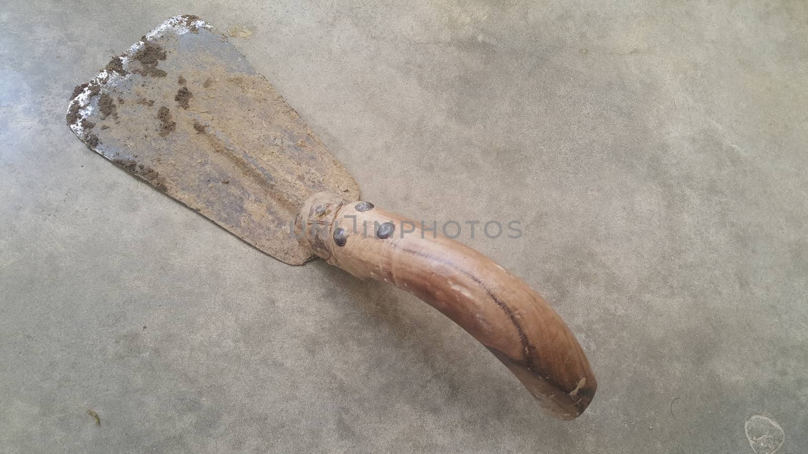 Closeup of old style garden trowel with metallic blade and wooden handle placed over a concrete floor. Trowel is old, rugged and dirty with mud.