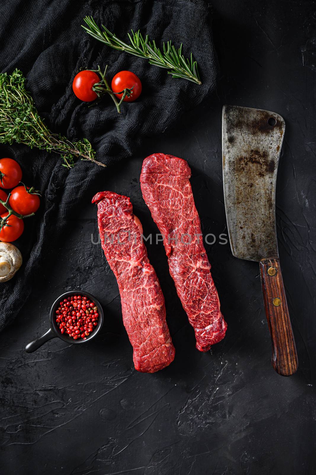 Raw set of denver cut organic meat with vegetable rosemary and other ingredients near butcher meat clever knife for bbq or grill over black stone background top view vertical.