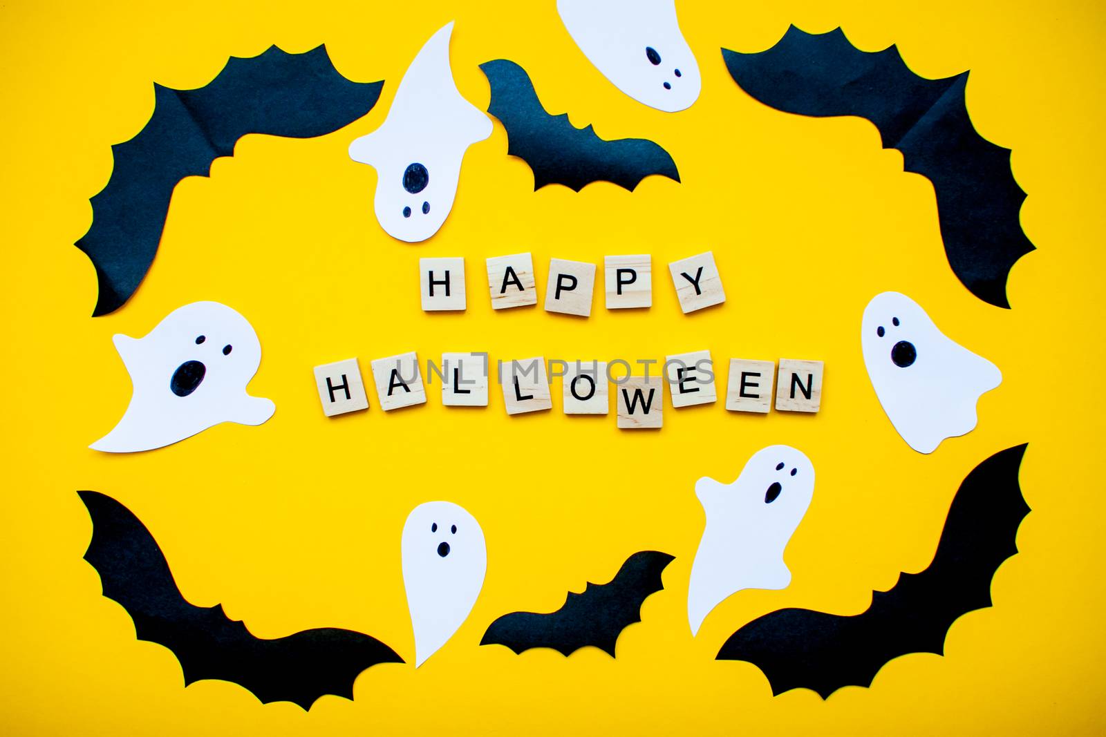 inscription from wooden blocks Happy Halloween and frame made of paper homemade bats and paper ghosts on a bright yellow background