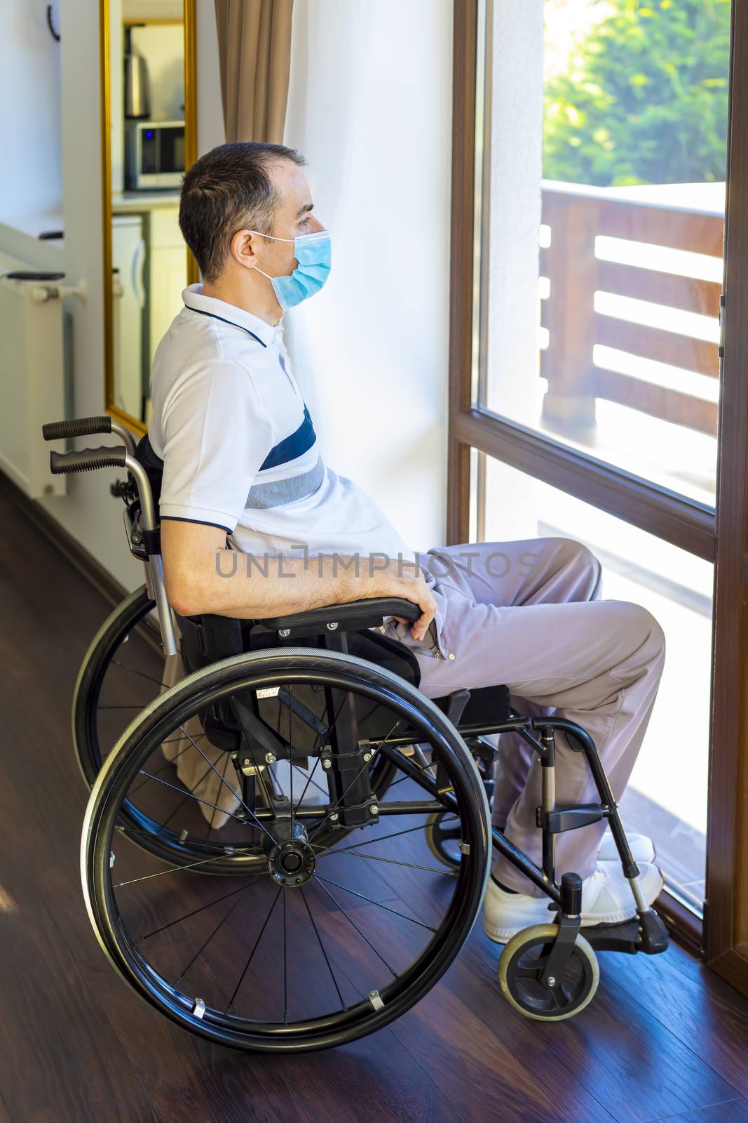 Lonely young man wearing face mask sitting in a wheelchair alone looking out the window. Focus on his face.