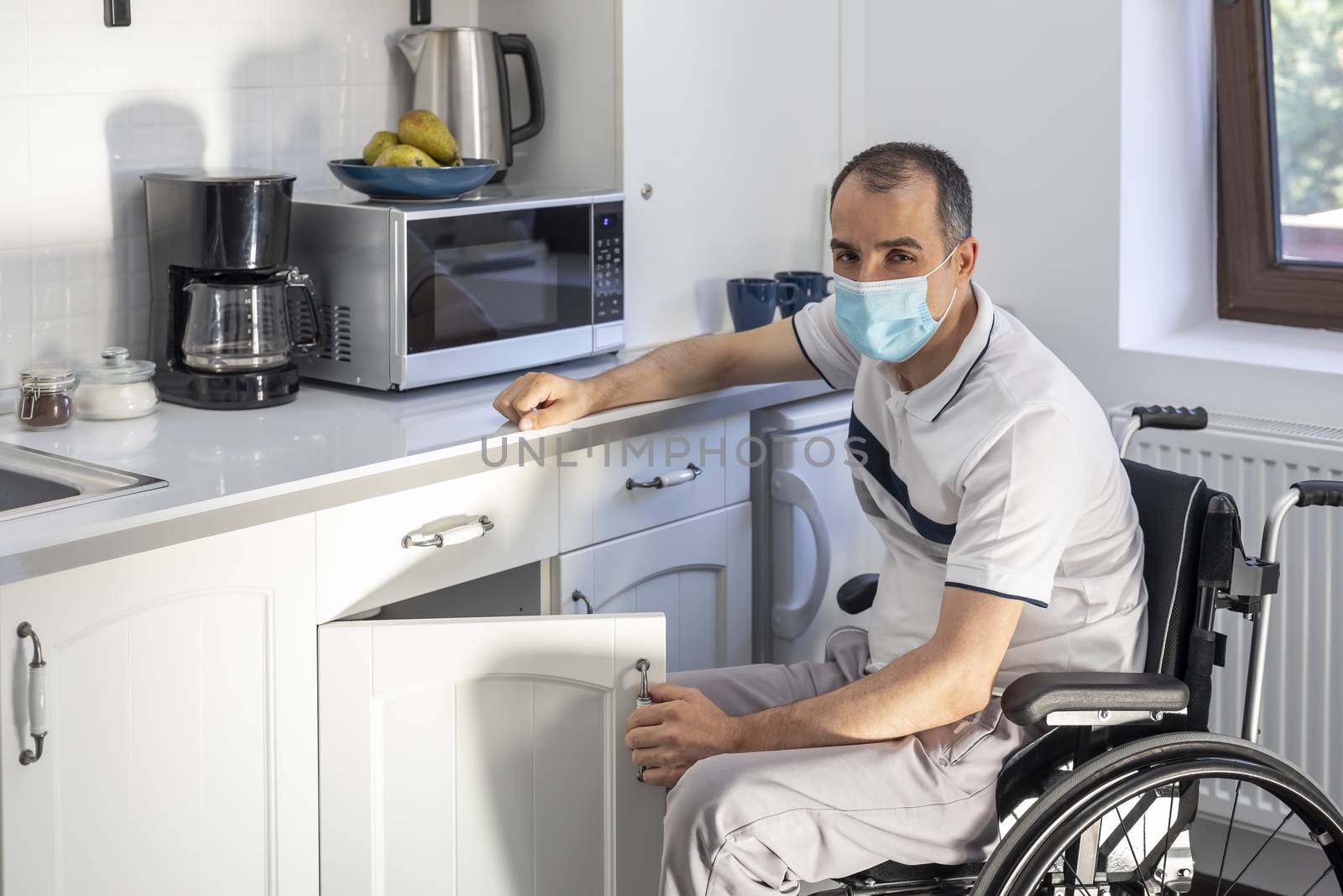 Smiling Young Handicapped Man Sitting On Wheelchair In Kitchen.  Young man wearing face mask sitting in front of kitchen. Focus on his face.
