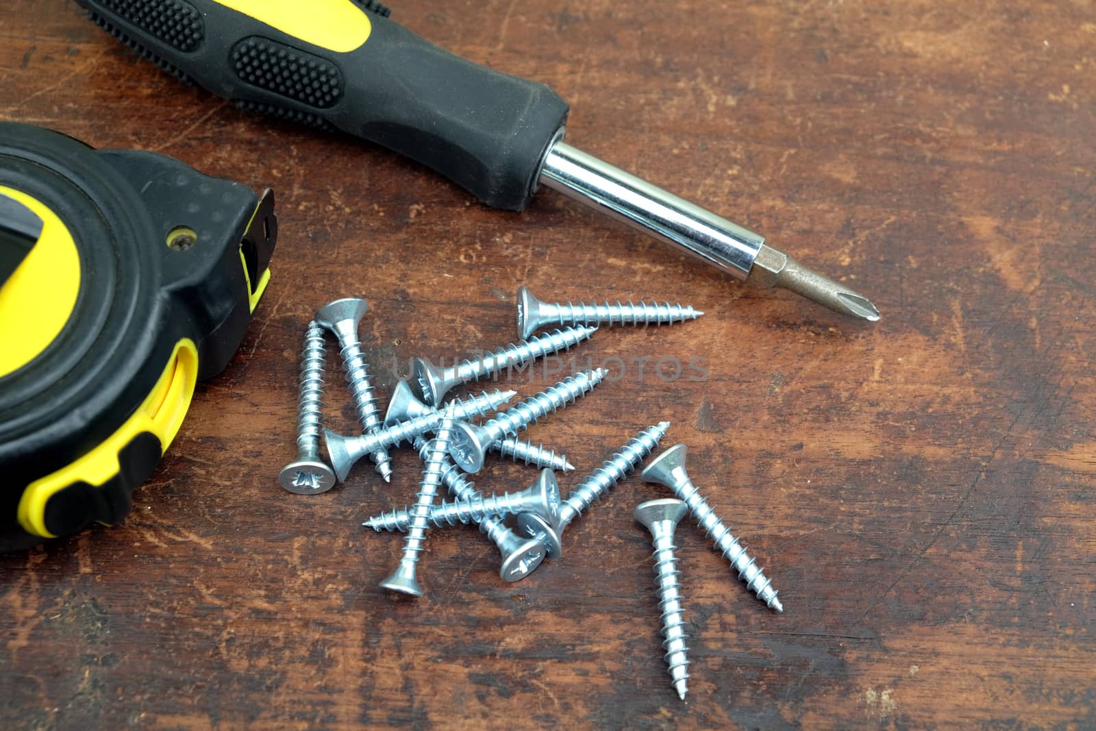 Heap of many light gray screws, screw driver and on brown wooden background closeup view