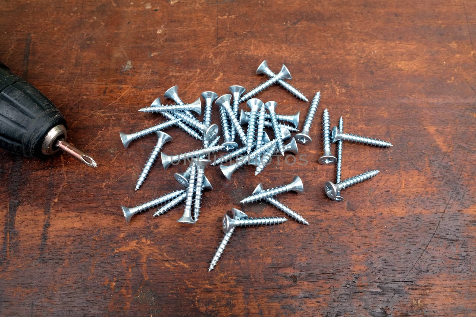 Heap of many light gray screws and old motor screw driver on brown wooden background top view by dymaxfoto