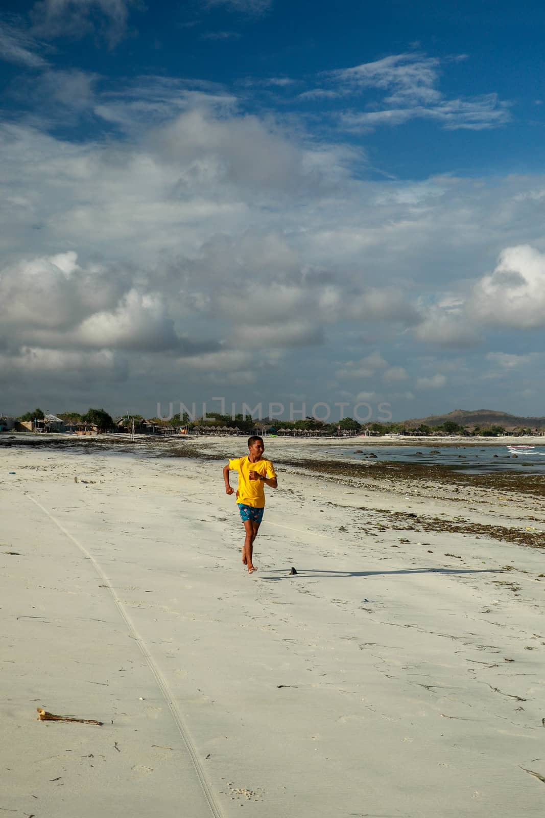 Running man jogging on beach. Indonesian teenager in shorts and a yellow T-shirt running along the beach. Young athlete trains by the sea. Fit young male sport fitness model exercising in summer.