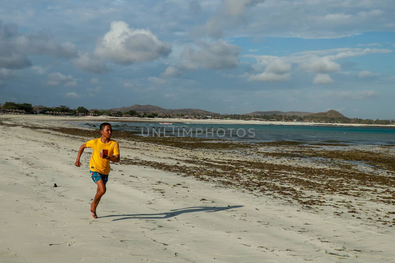 Running man jogging on beach. Indonesian teenager in shorts and a yellow T-shirt running along the beach. Young athlete trains by the sea. Fit young male sport fitness model exercising in summer.