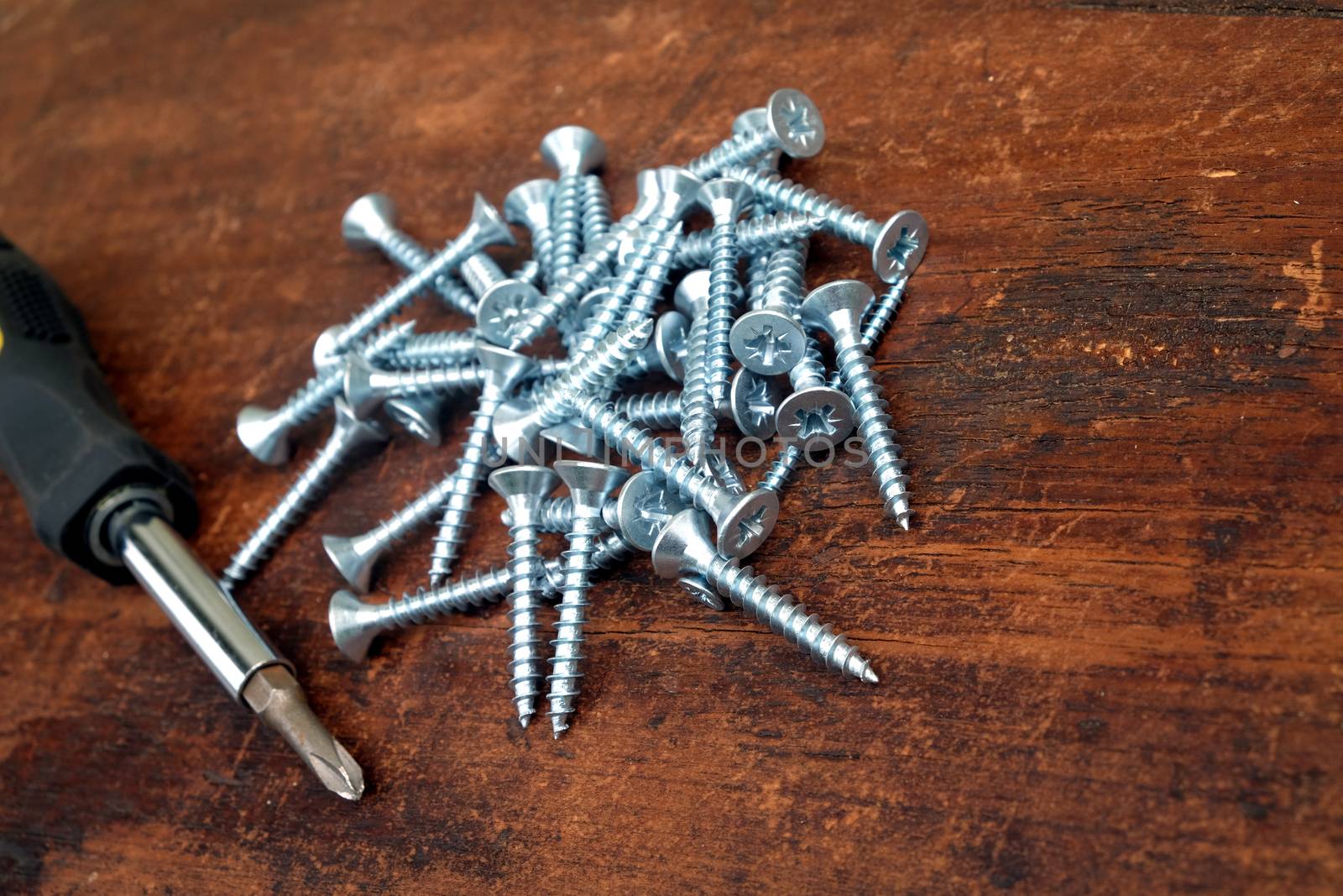 Heap of many light gray screws and old screw driver on brown wooden background closeup view