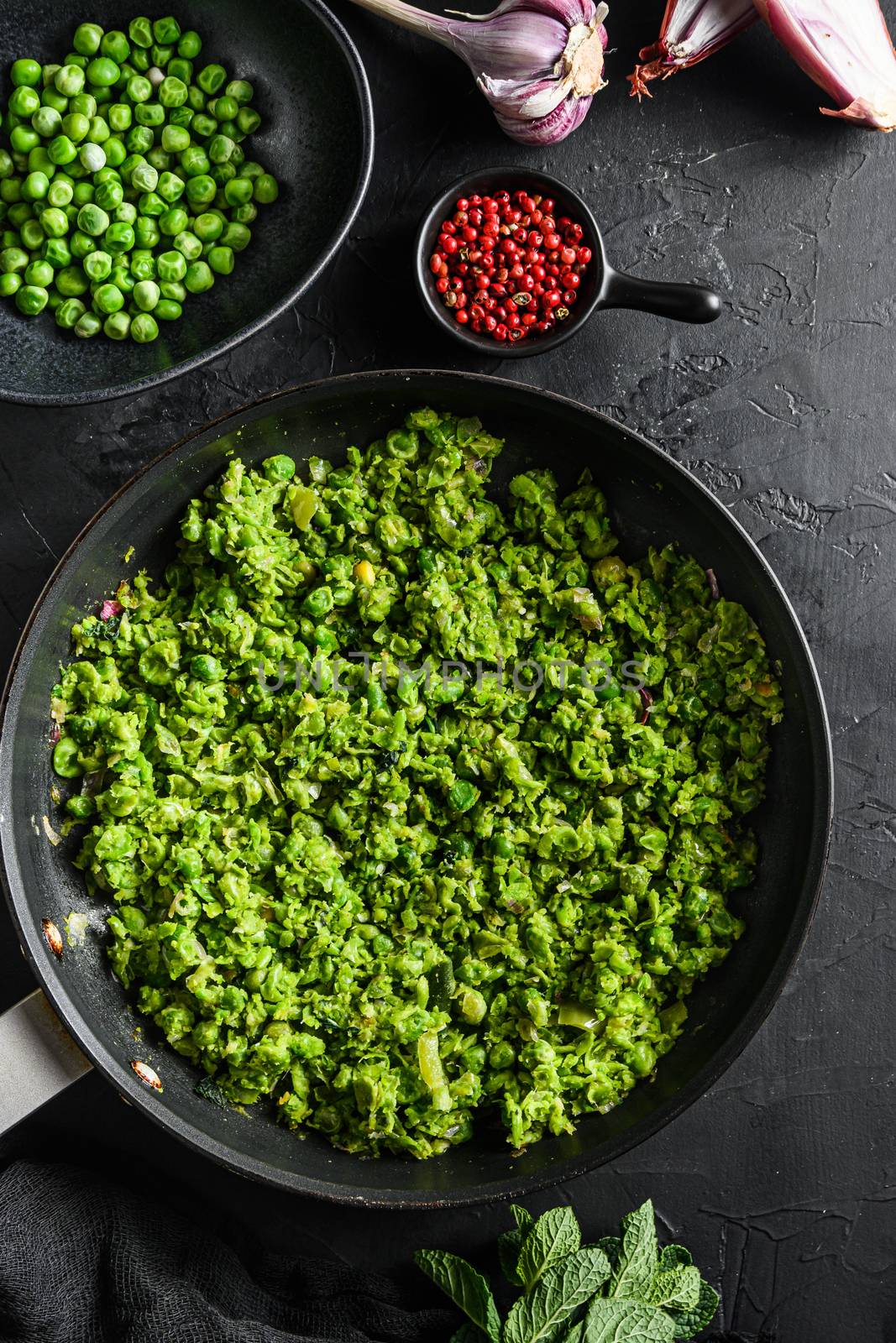 Mushy peas recipe cooked frying pan and peas in bowl with mint shallot pepper and salt on black stone surface organic keto food top view close up.