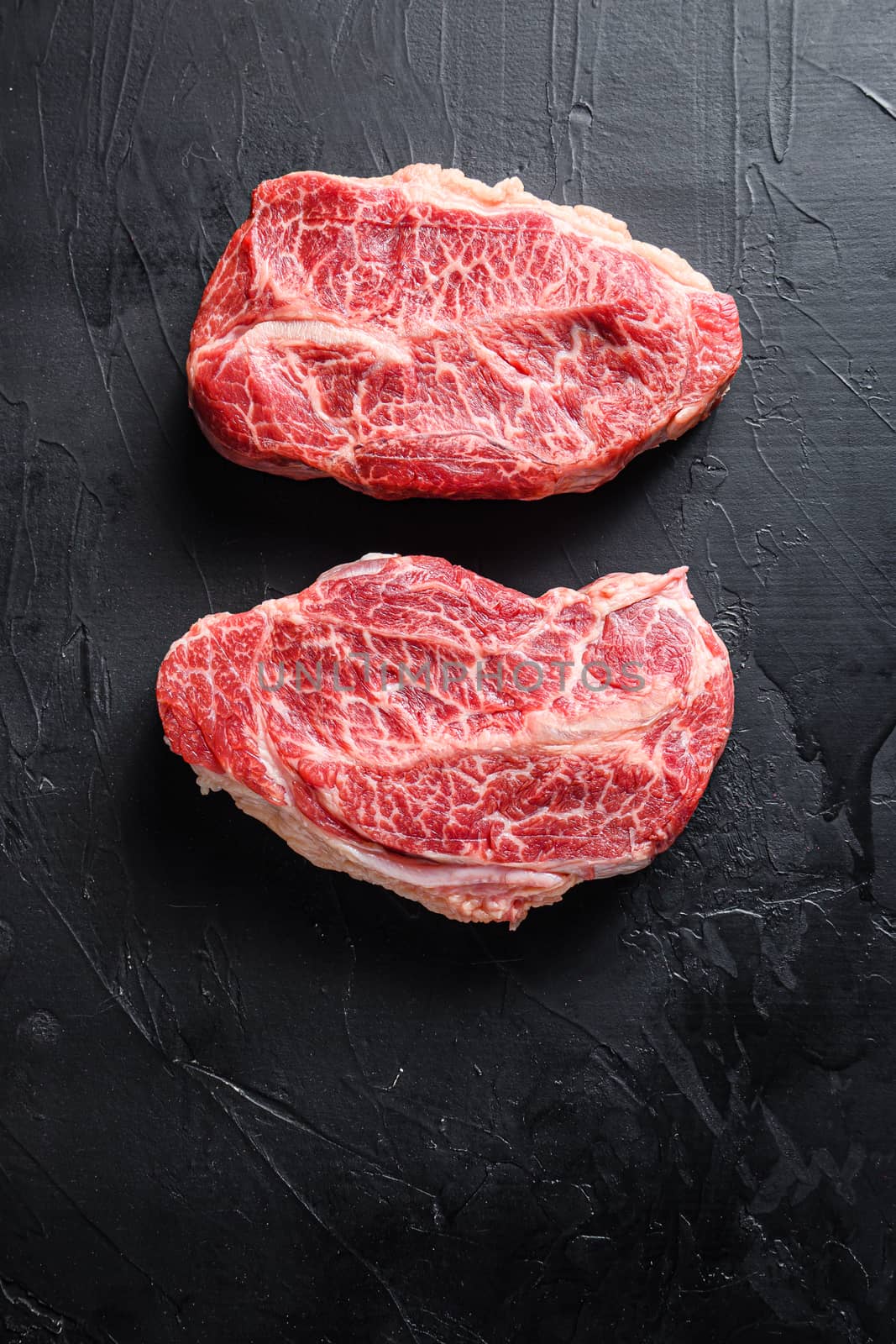 Raw top blade flat Ironcut, on black slate , bio marbled beef with herbs tomatoes peppercorns overblack stone surface background top view close up.