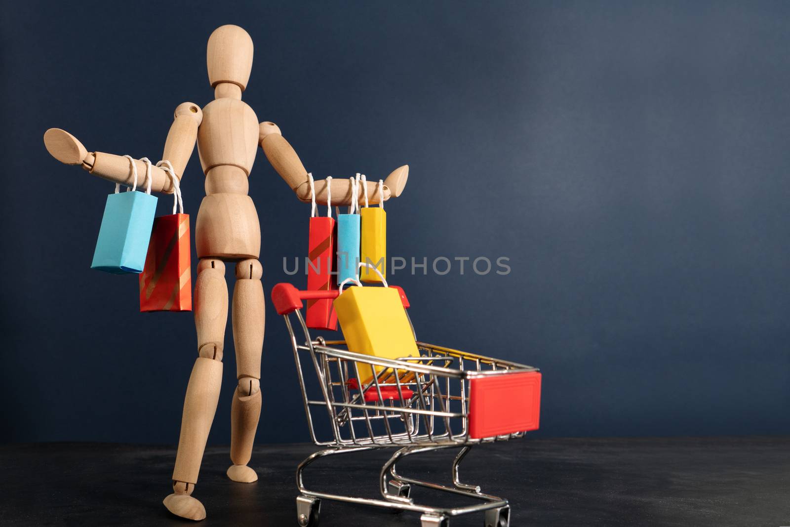 Cheerful happy shopaholic wooden doll with lots of shopping bags on arm and shopping cart