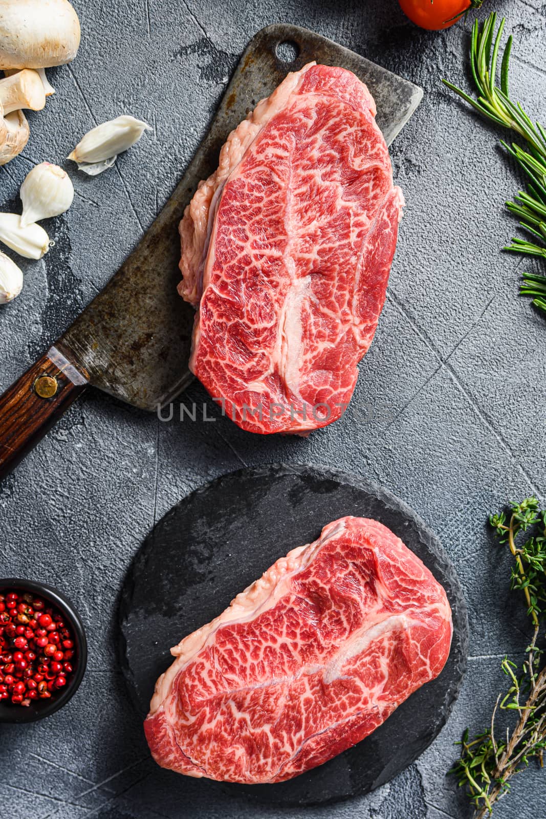 Raw top blade flat Ironcut, on black slate , and meat butcher cleaver marbled beef with herbs tomatoes peppercorns over grey stone surface background top view close up.