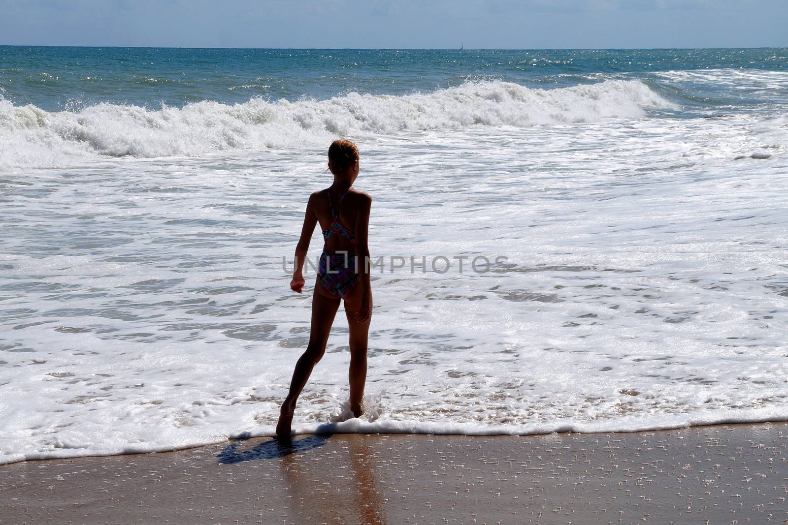 teenage girl walking alone along the empty seashore on the sunny day, rear view.