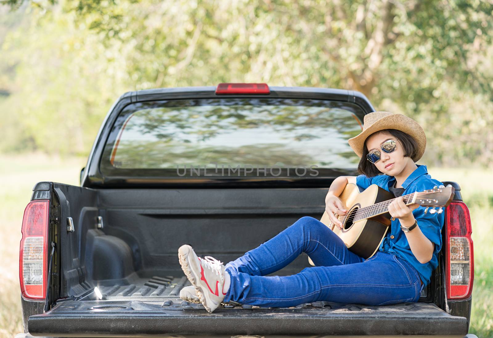 Young asian women short hair wear hat and sunglasses playing guitar ,sit on pickup truck in countryside Thailand