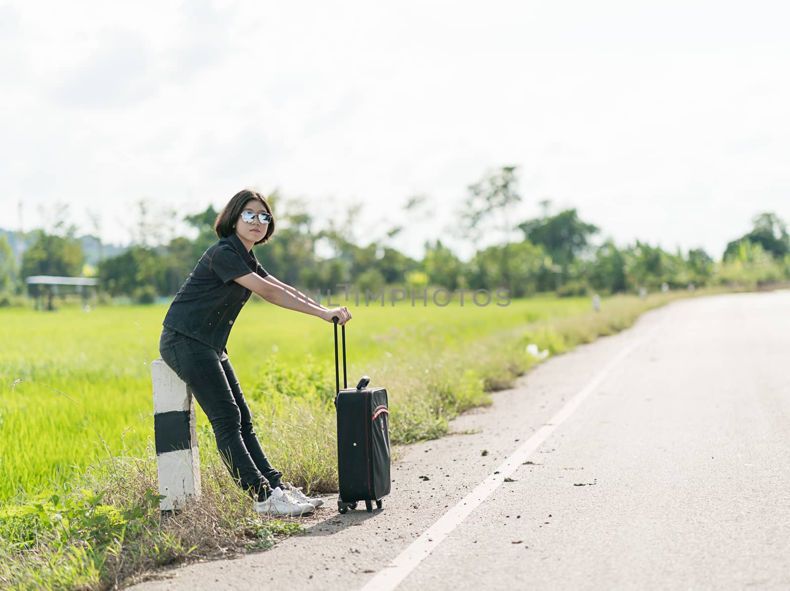 Young asian woman short hair and wearing sunglasses with luggage hitchhiking along a road in countryside Thailand