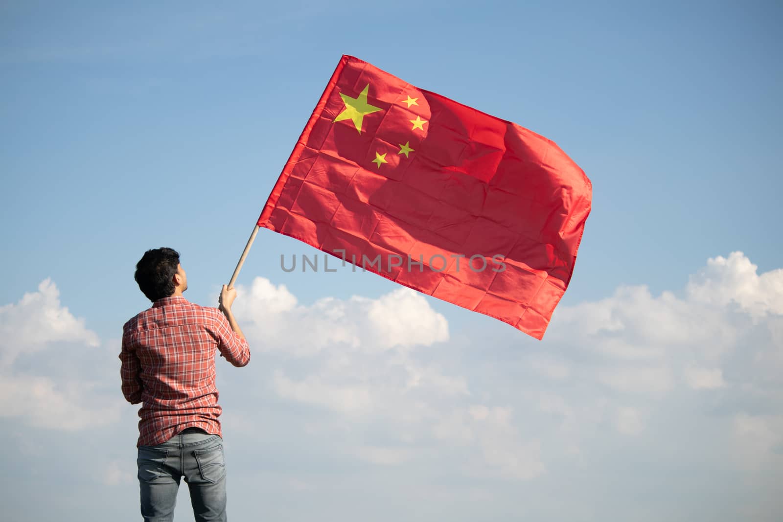 Young man proudly holding waving Chinese flag on top of mountain peak - Concept showing Celebration of Chinese Republic or national Day by lakshmiprasad.maski@gmai.com