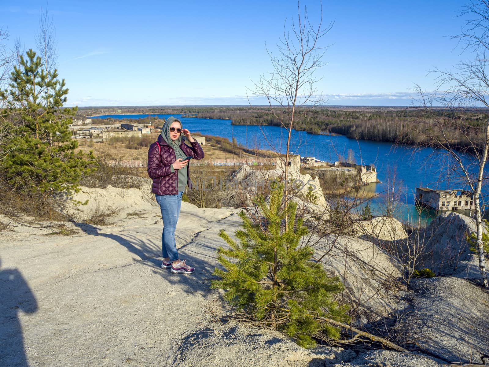 Hiker on mountain top with smartphone. Woman is standing and taking a picture on smartphone. Abandoned Quarry Of Rummu, Estonia. Scenic View Of Land Against Clear Blue Sky. Panoramic View.