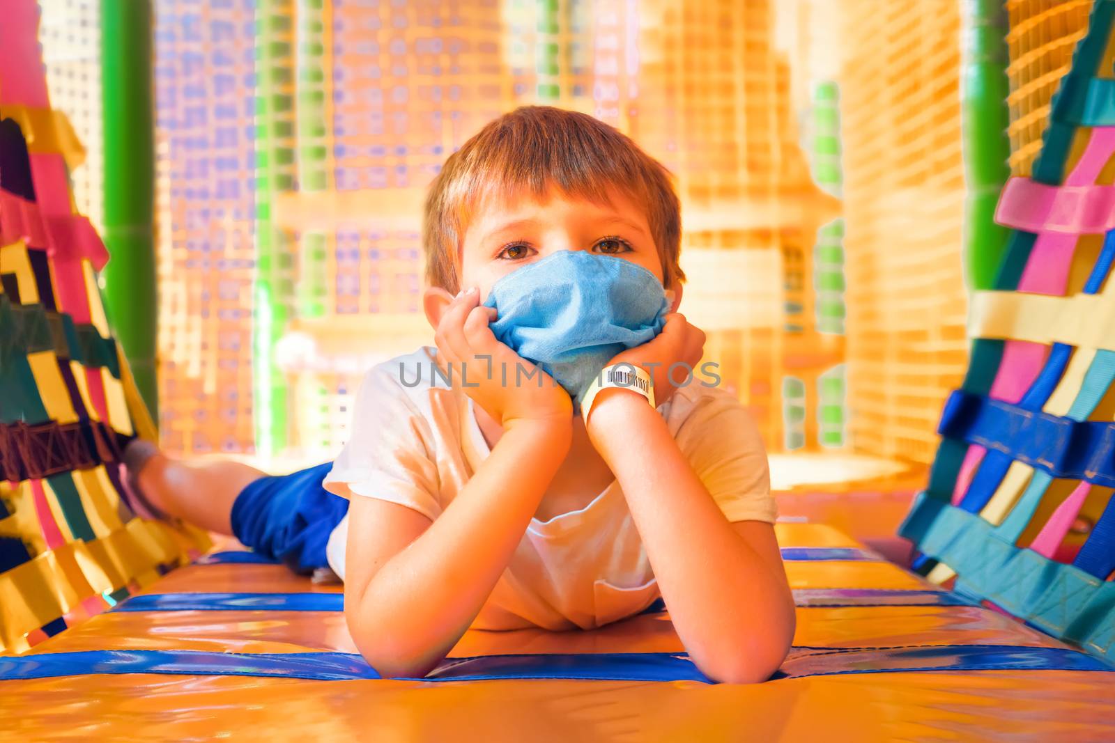 Schoolboy in protective mask lying on the floor in playing room. Elementary school during quarantine. Cheerful boy in mask in kindergarten. Social distancing, preventing illness. Childhood, quarantine by synel