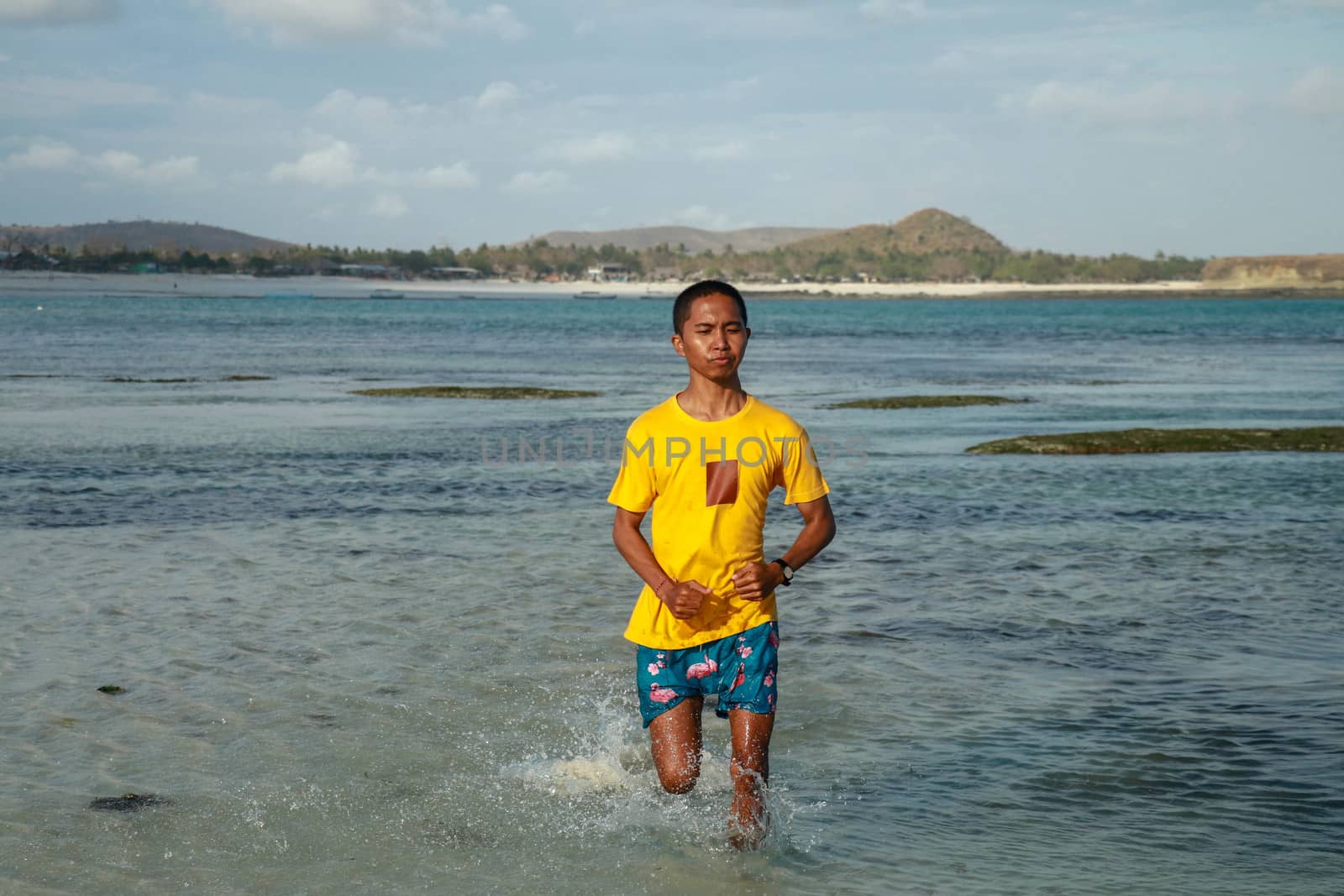 Topless, athletic and healthy black man running along the beach, splashing water during sunset. Teenager in a yellow T-shirt running in shallow water. Young man in shorts trains on the beach.