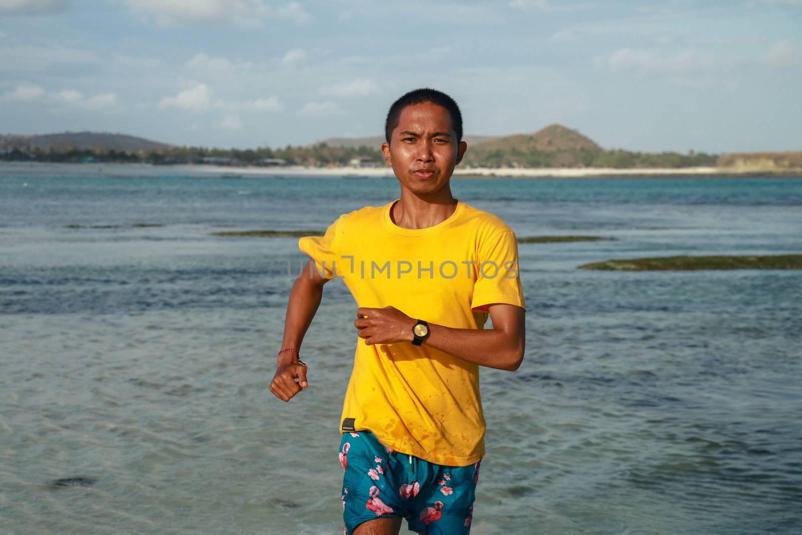 Portrait of a smiling handsome man running and enjoying the view of the ocean. Close up of happy teenager in yellow t-shirt running in shallow water. Youg asian man in shorts trains on the beach.