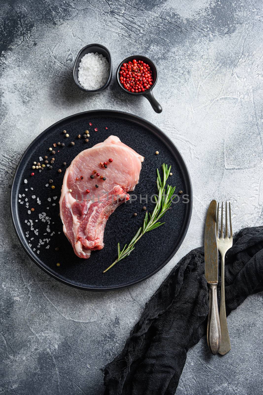 pork meat, cutlet in a black round plate on a grey textured stone background with fork and knife rosemary garlic peppercorns ingredients top view vertical by Ilianesolenyi