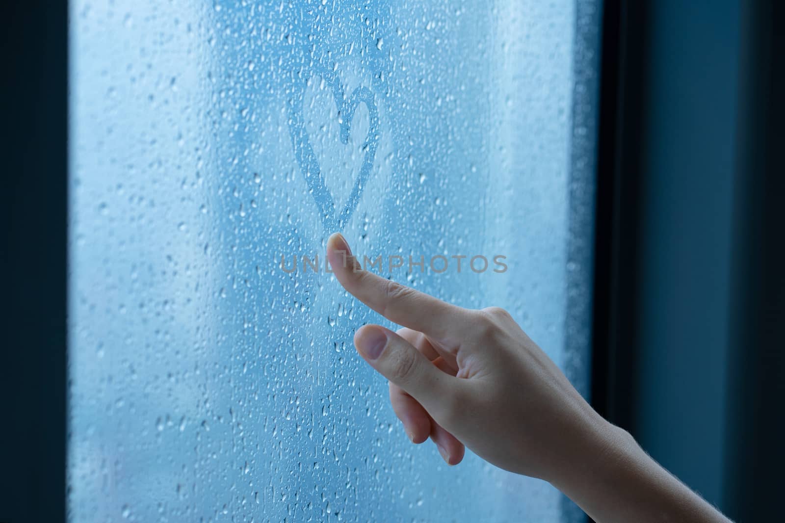 Female hand drawings a heart on a foggy window during the rain. Glass in drops of water. The concept of romance
