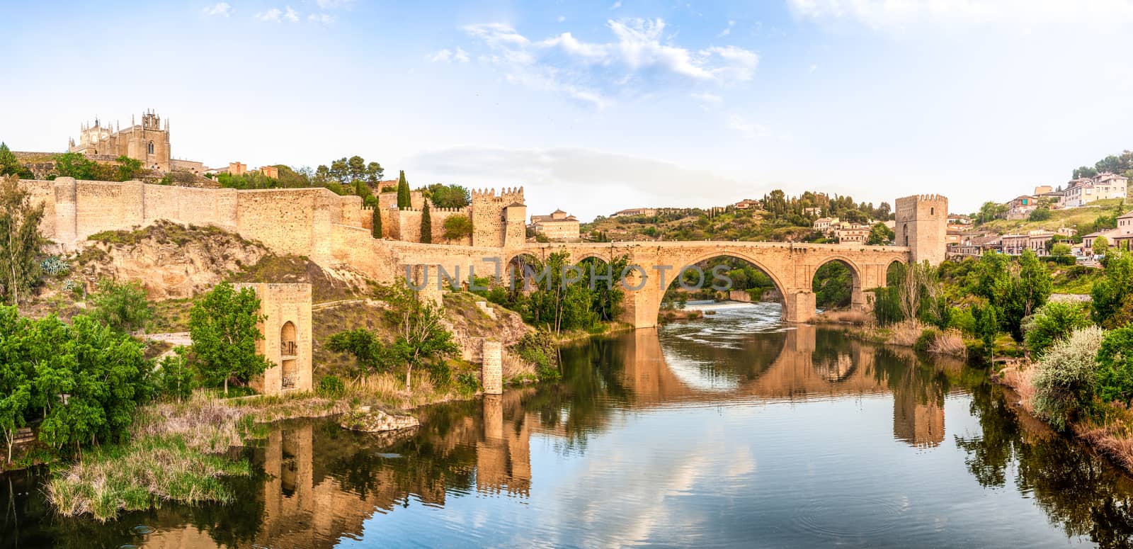 Beautiful landscape of Toledo in Spain. Stone bridge across calm river. Blue sky reflected in crystal clear water. Big fort and country houses in background. Popular tourist place in Europe.