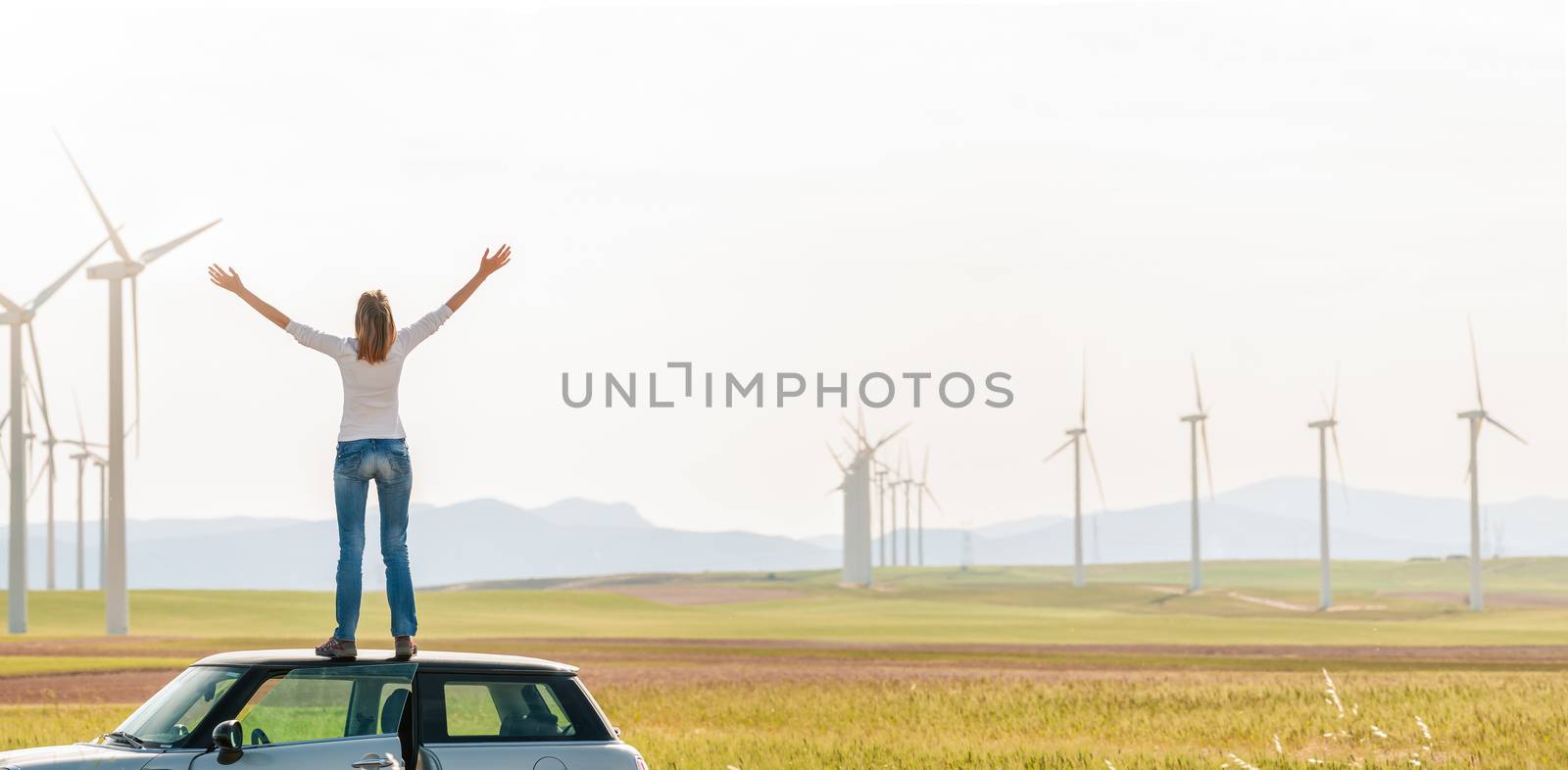 Young woman standing on top of her car with hands raised. Girl in yellow fall field with wind turbines in background. Alternative energy source. Power generation. Ecology and freedom concept.