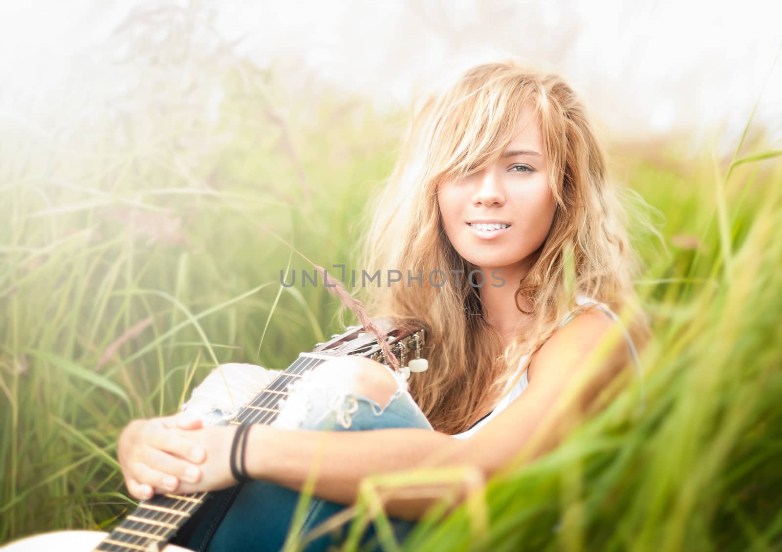 Pretty girl with guitar sitting on ground. Beautiful woman with blonde hair holding musical instrument and smiling. Green grass as background. Sunny summer day. Outdoor activity for young people.