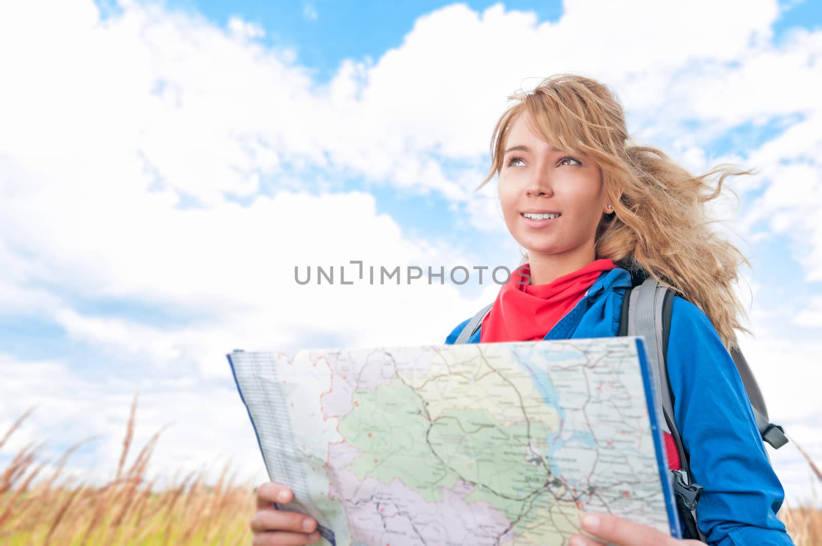 Young pretty woman tourist standing in wheat field with map. Blue cloudy sky in background behind girl. Tourism travel and hiking outdoor in summer. Healthy lifestyle.
