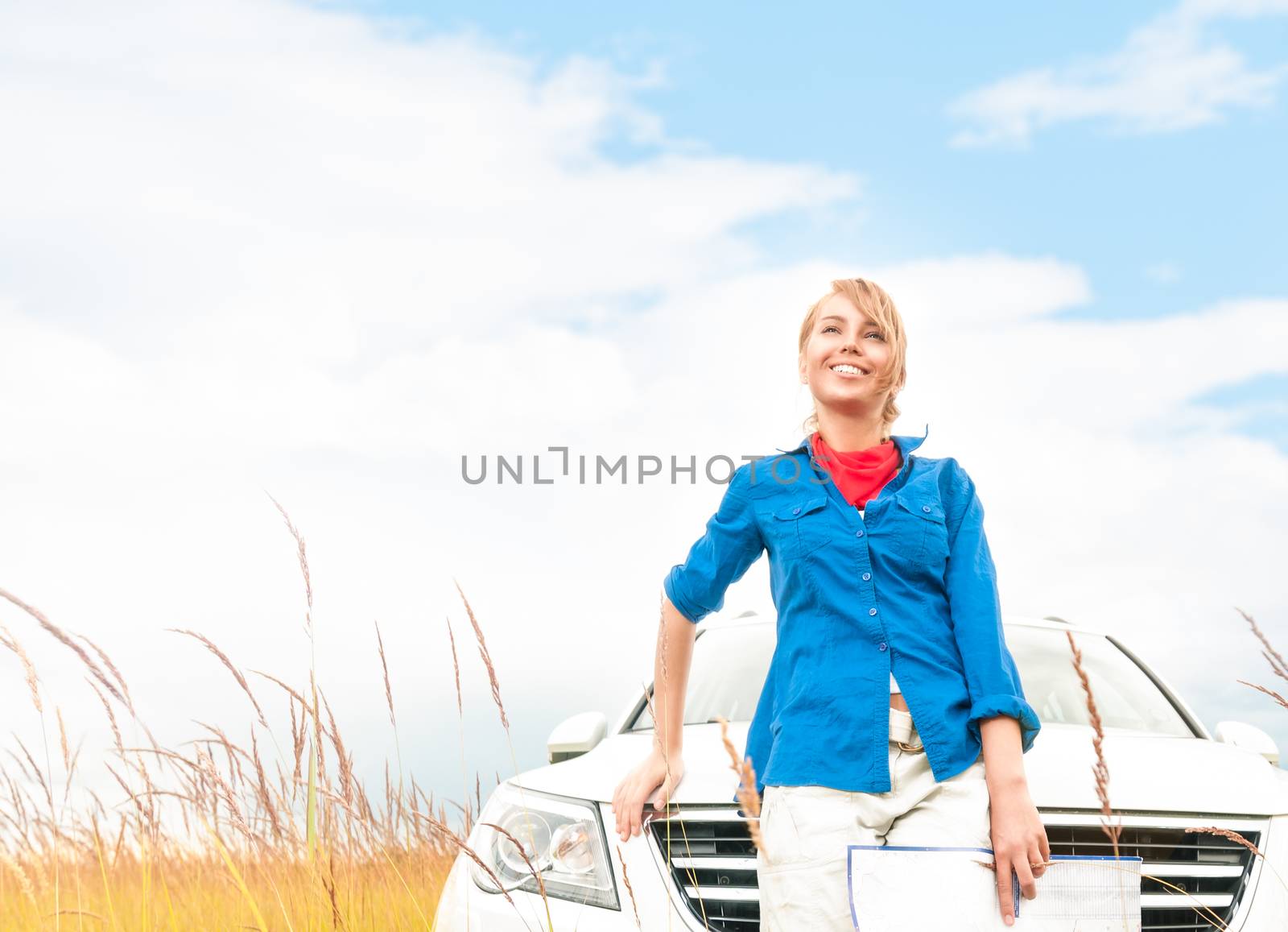 Young pretty female tourist with map in front of car. Cloudy sky and wheat field background. Happy and smiling girl looking far away. Tourism and travelling by car in summer and fall.