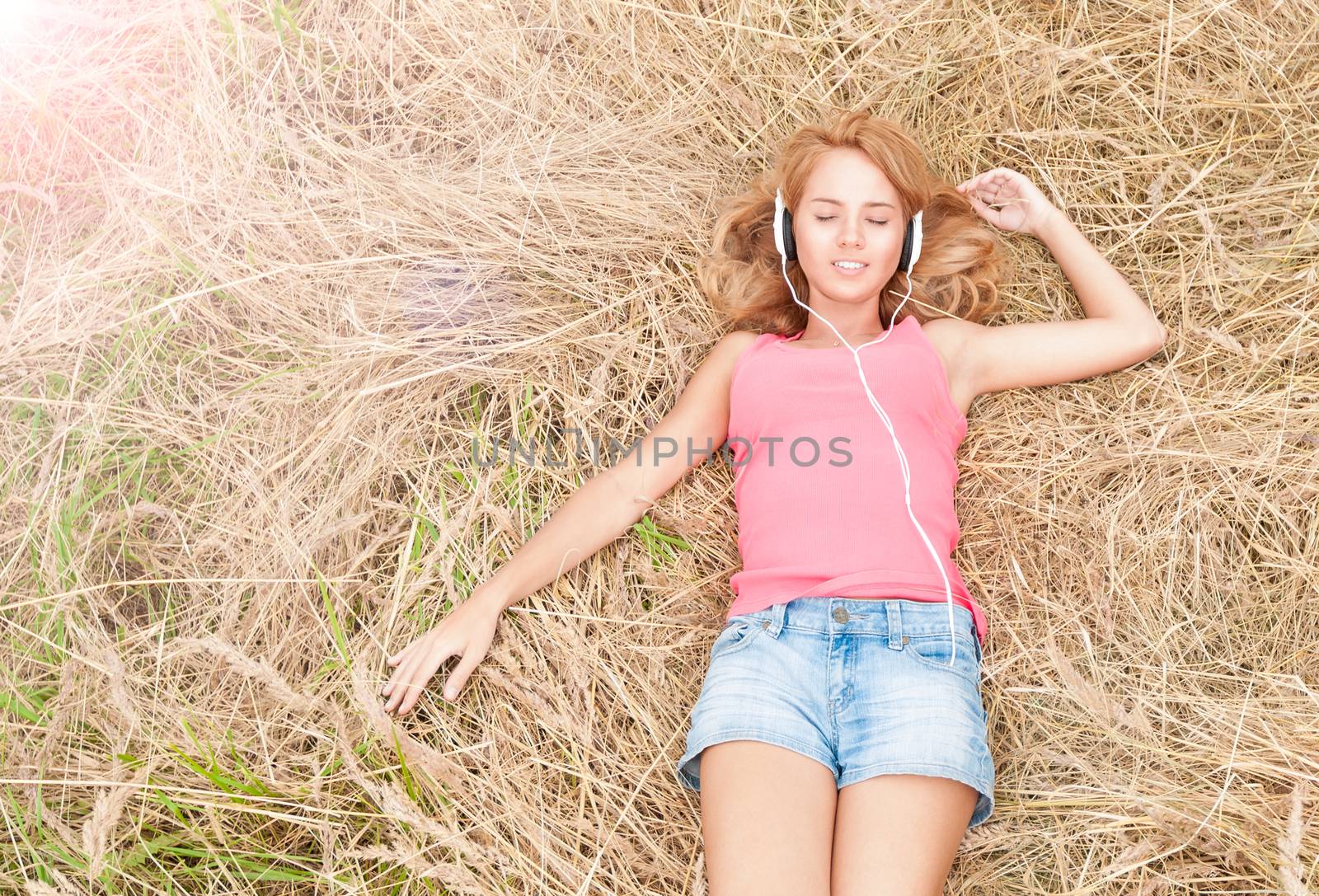 Beautiful girl relaxing in headphones outdoors. Pretty smiling woman with closed eyes listening to music lying on hay in field. Harmony of human and nature. Countryside.