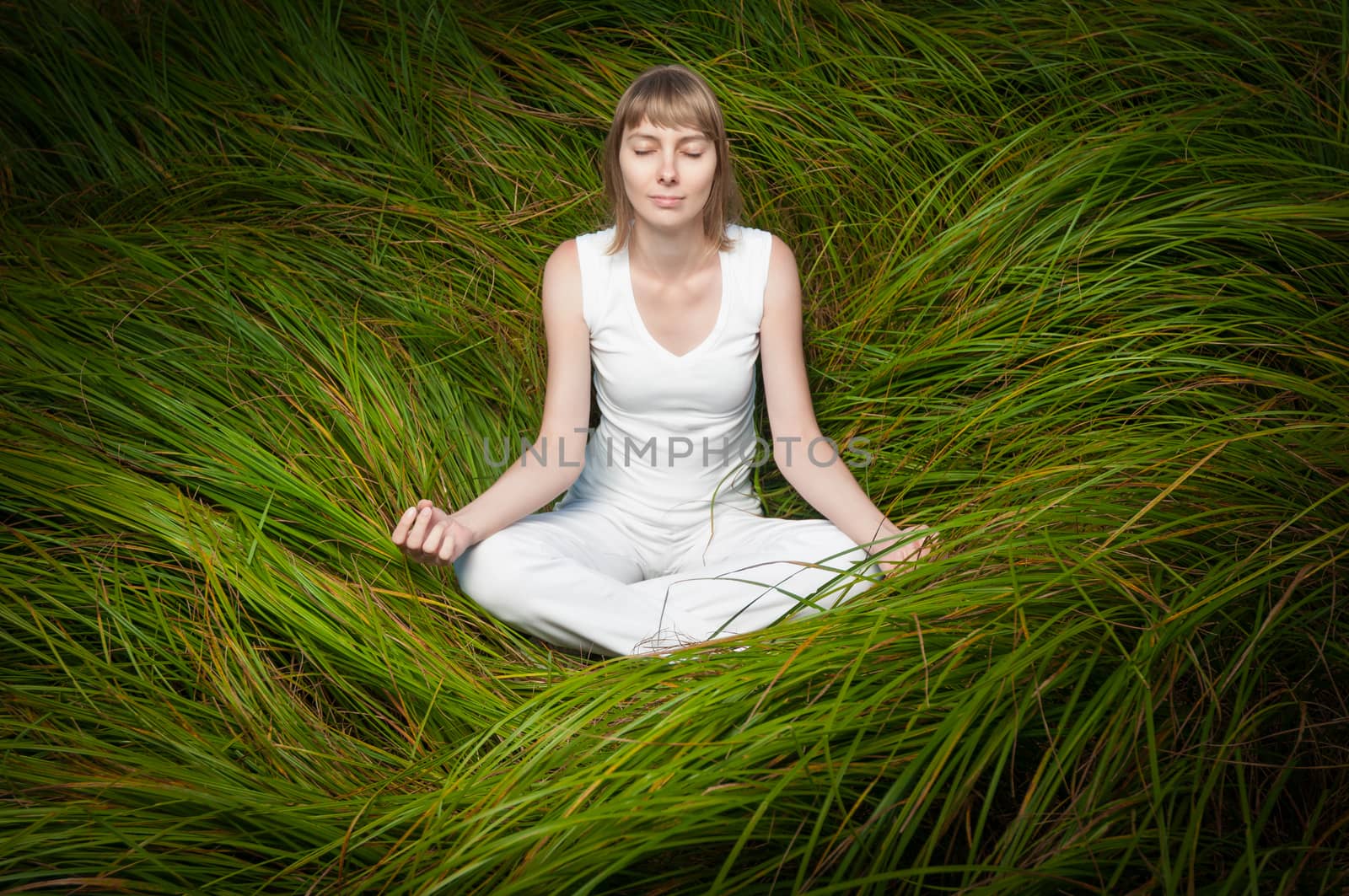 Cute blonde girl in white clothes meditating on green grass. Woman sitting in lotus position, eyes are closed, her hands lay on her knees. Girl is alone. Atmosphere of unity with nature and harmony.