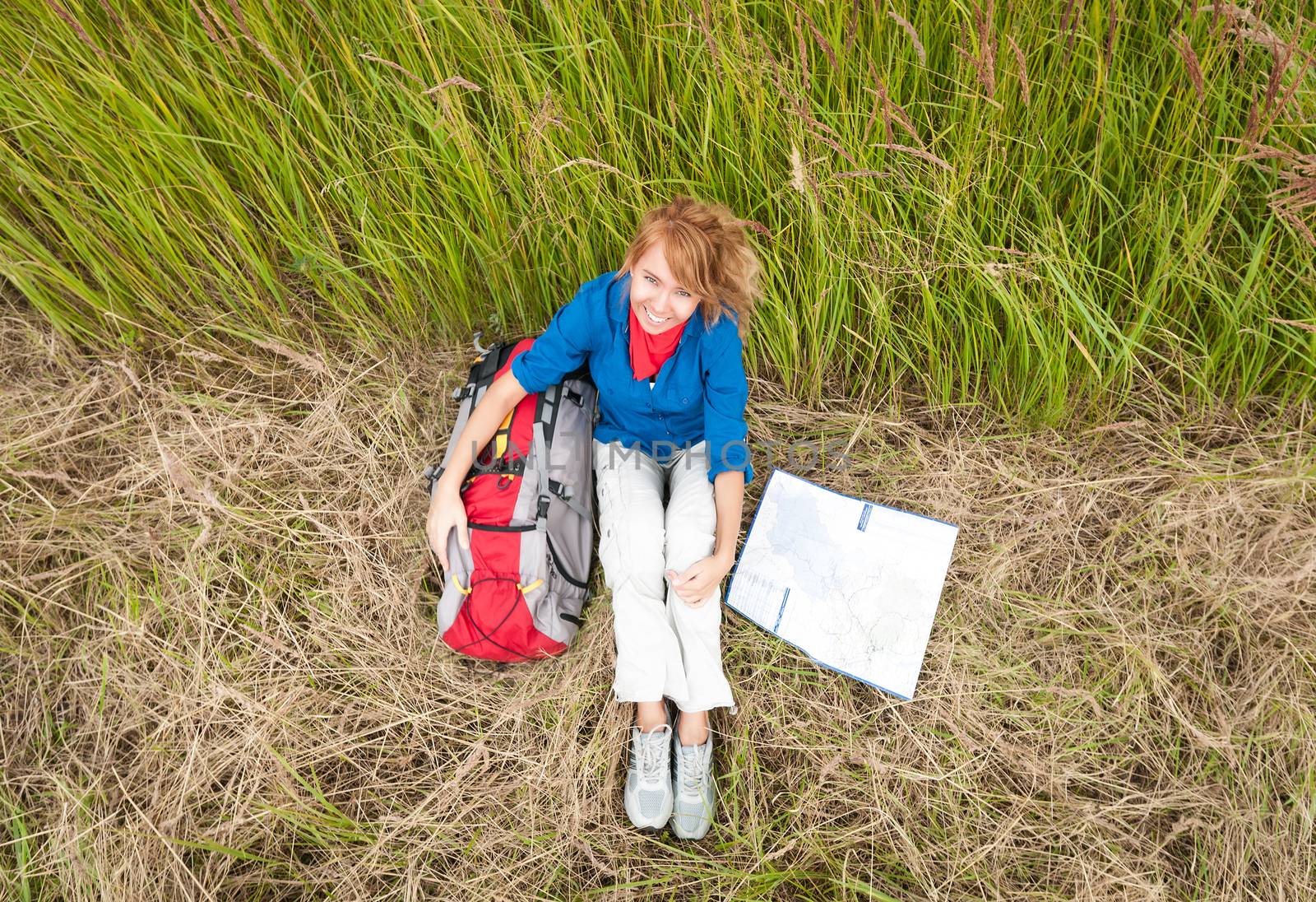 Young pretty female tourist with backpack and map having stop in field. Happy and smiling girl on halt sitting on dry grass. Tourism travelling and hiking outdoors in summer.Healthy lifestyle.