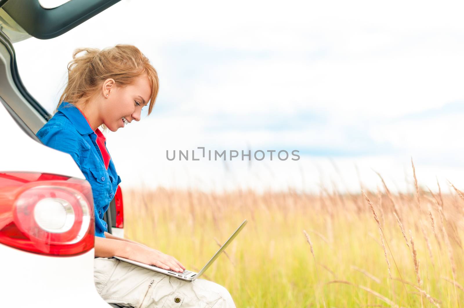 Young happy girl sitting and working on computer. Beautiful woman near car in meadow holding laptop. Person working outdoor. Tail light of car in foreground and field with sky in background.