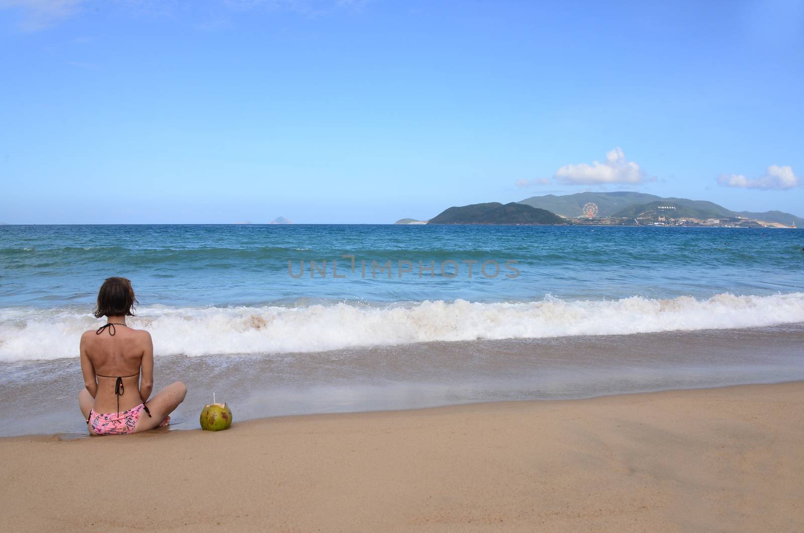 NHA TRANG, VIETNAM – 29 FEBRUARY 2020 : Sexy bikini body woman sitting on nha trang  beach behind is an Vinpearl Land