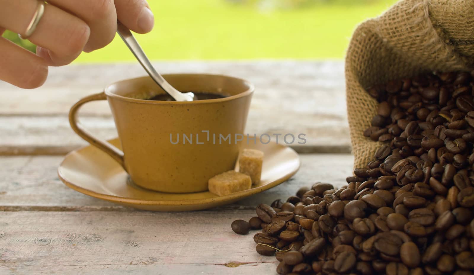 Close-up of coffee beans in a burlap jute sack and cup of coffee on a wooden table outdoors. Hand stirring sugar in a cup