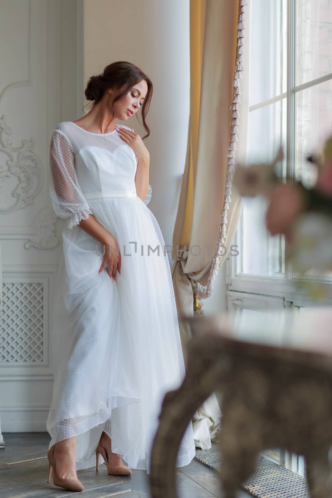 Full-length portrait of the bride in a white wedding dress by the window.