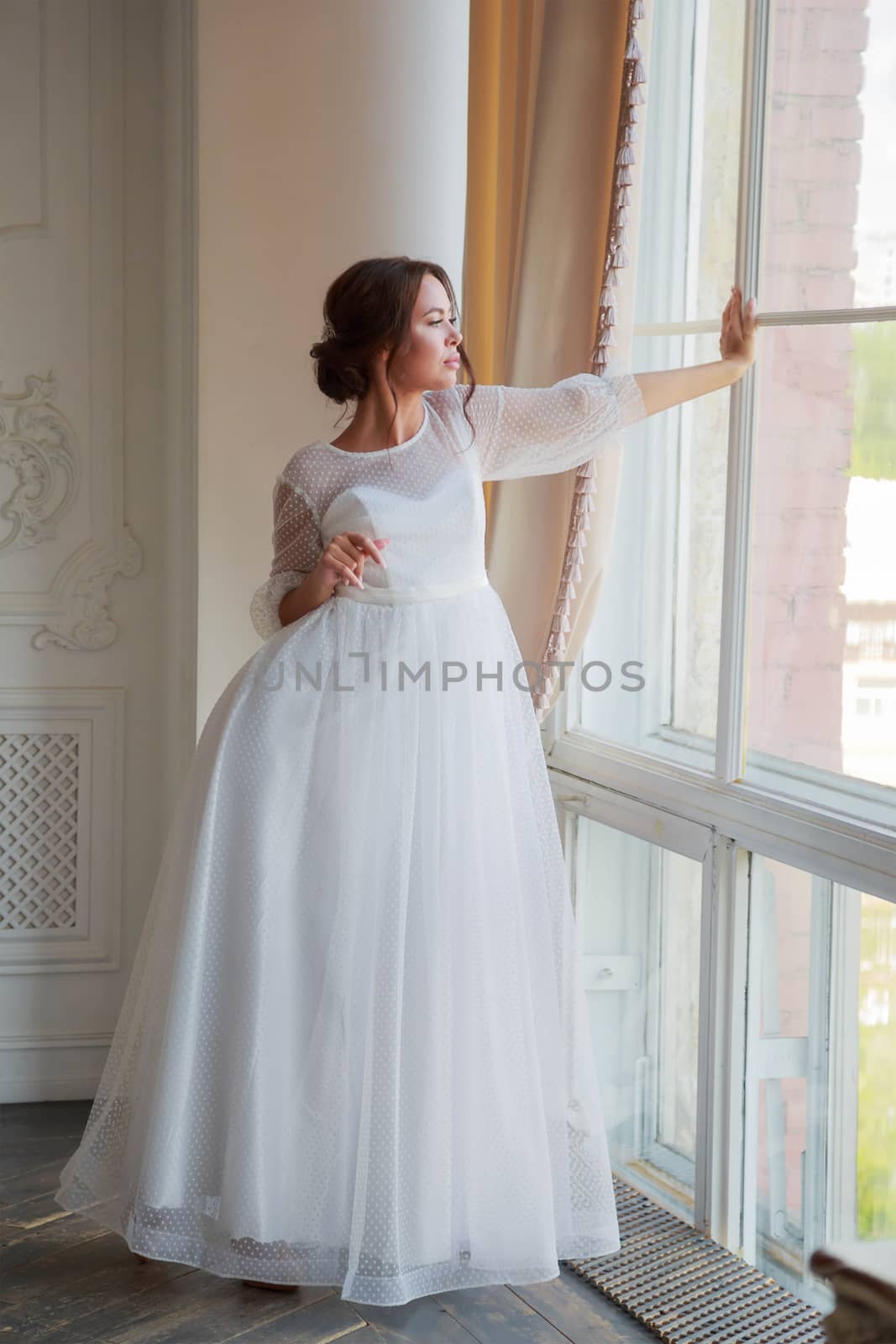 Full-length portrait of the bride in a white wedding dress by the window.