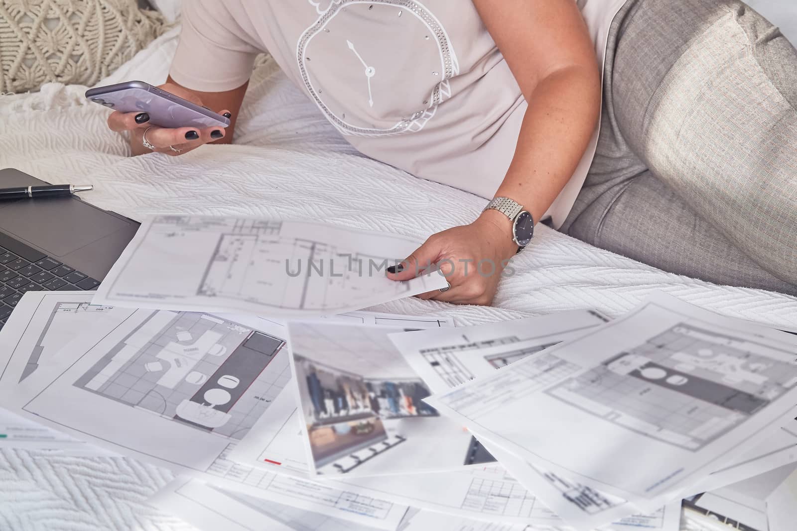Woman interior designer lies on the bed with interior plans for a new project and notebook, holds a mobile phone in her hands, freelancer talks with a customer. Soft focus