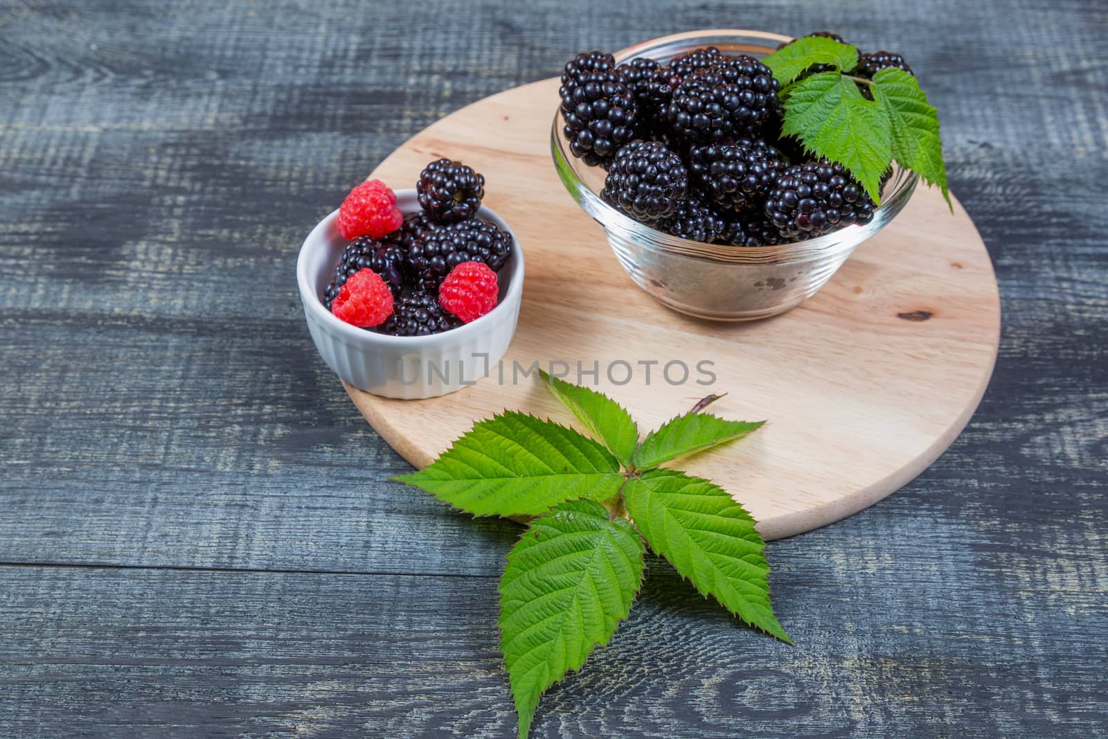 ripe blackberry with leaves on a wooden cutting board in a glass bowl on dark blue wooden background.